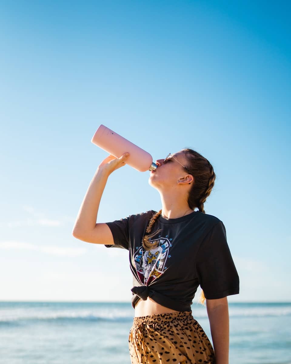 woman drinking water using pink bottle beside sea during daytime